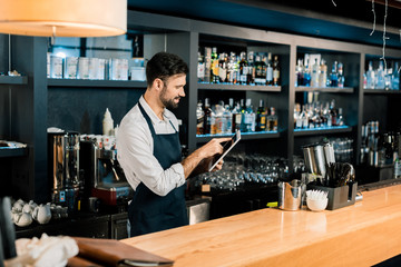 Barman standing in apron and typing on digital tablet
