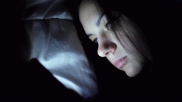 Young woman lying under blanket in the night and typing on mobile phone in bed in close-up