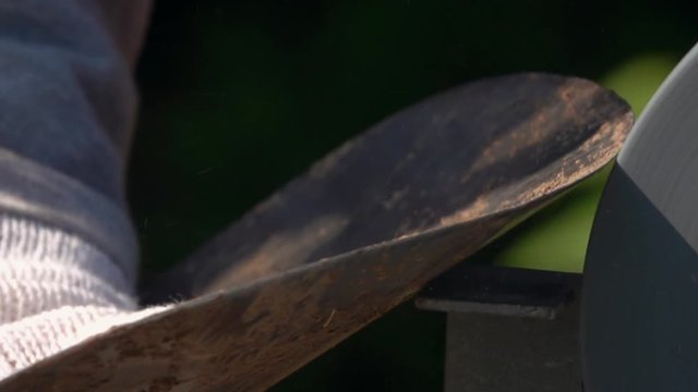Sharpening of soiled garden tool with unbalanced grinding machine in slow motion. Panning of machinery scene with round-pointed steel shovel, waving hand in safety glove and turning grindstone wheel.