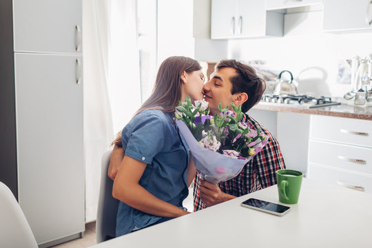 Young man gifting bouquet of flowers to his girlfriend in kitchen. Happy couple hugging. Romantic surprise