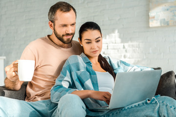 Fototapeta premium handsome husband holding cup of coffee while wife using laptop on couch