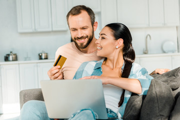 handsome husband holding credit card while smiling wife using laptop and doing online shopping at home