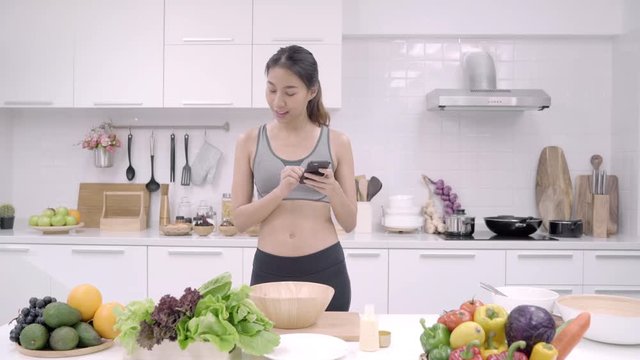Young Asian woman making salad healthy food while using mobile phone for looking recipe in the kitchen, beautiful female use vegetables preparing salad for fit body at home. Healthy food concept.