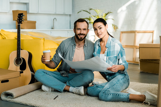 Handsome Husband And Wife Drinking Coffee And Holding Blueprint Of New House, Moving Concept