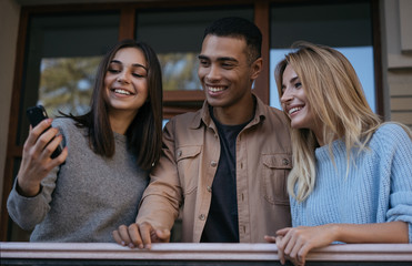 Happy multiracial hipsters with emotional smiling faces using smartphone for mobile photo, they taking selfies. Positive stylish friends using mobile application, shopping online, booking tickets
