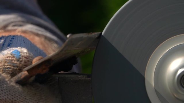 Sharpening of grange garden tool with unbalanced grinding machine in slow motion close up. Panning of traditional machinery scene with detailed hoe, waving hand in safety glove and turning grindstone.