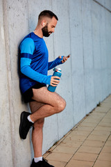 Male runner leaning on wall and using smart phone. In other hand bottle with water.