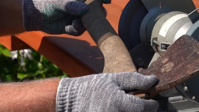 Sharpening of old grange ax with unbalanced grinding machine in slow motion. Panning of machinery scene with retro tool, waving man hands in safety gloves, turning grindstone wheel and bright sparks.