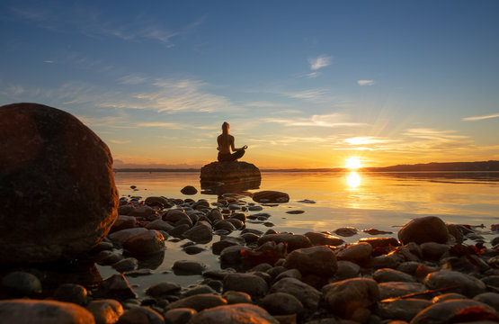 h&uuml;bsche junge frau macht yoga auf einem Stein im Wasser See Meer im Sonnenuntergang Abendrot