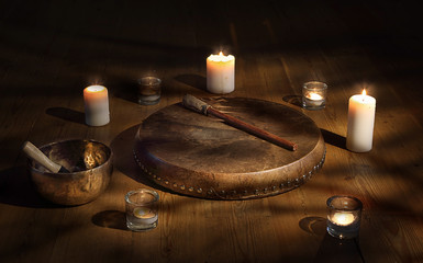 Shaman tambourine and Tibetan bowl surrounded by candles in a dark room.
