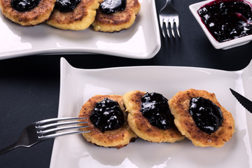 2 white stylish plates with cottage chesse pancakes, metal cutlery and jam. Dessert on dark background.