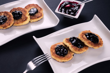 2 white stylish plates with cottage chesse pancakes,  fork and jam. Dessert on dark background.