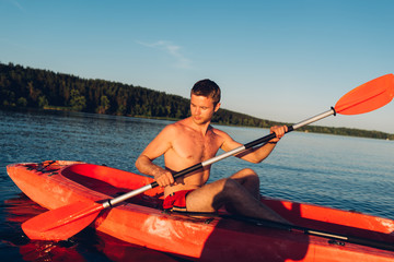 young guy without t-shirt is swimming on a red kayak with a paddle in his hands, close-up