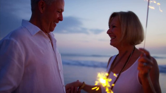 Retired Caucasian couple enjoying sunset with sparklers on beach vacation