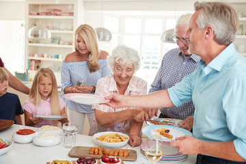 Multi-Generation Family And Friends Eating Food In Kitchen At Celebration Party