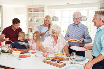 Multi-Generation Family And Friends Eating Food In Kitchen At Celebration Party
