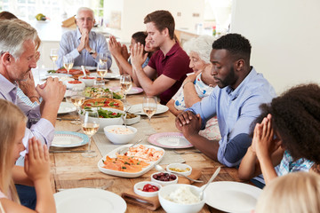 Multi-Generation Family And Friends Around Table Praying Before Meal At Party