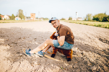 Adult stylish man riding wooden toy horse on field outdoor. Young funny unusual strange male person with smiling face lifestyle portrait. Odd happy boy childhood memories. Country happy guy relaxing.