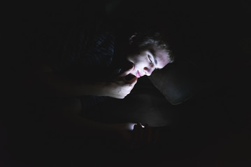 Curious teenager browsing the internet on his mobile phone as he is lying on a couch in the dark.