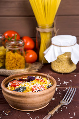 A clay plate with pasta with basil, tomato and parmesan next to a fork and cans of dry pasta on a wooden table