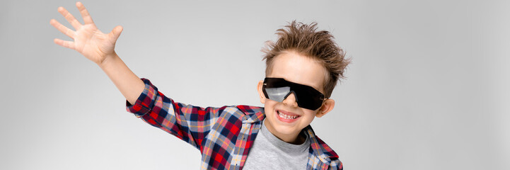 A handsome boy in a plaid shirt, gray shirt and jeans stands on a gray background. The boy in black sunglasses. The boy spread his hands to the sides.