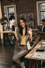 casual businesswoman holding smartphone with blank screen and showing thumbs up sign in loft office with colleagues working behind