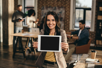 Fototapeta premium happy casual businesswoman holding tablet with blank screen in loft office with colleagues working behind