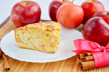 Appetizing piece of apple pie on a white plate next to cinnamon and red apples