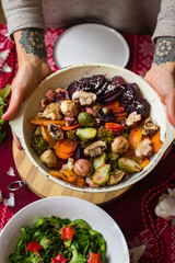 Woman hands hold skillet with baked, roasted, grilled vegetables. Beetroot, carrot, mushrooms, pumpkin, brussels sprouts with avocado dpi sauce. Vegan lunch, vegetarian Christmas food, red tablecloth