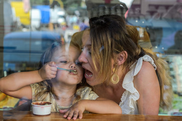 Young mother and daughter eat ice cream in a cafe, happy family, view through the window