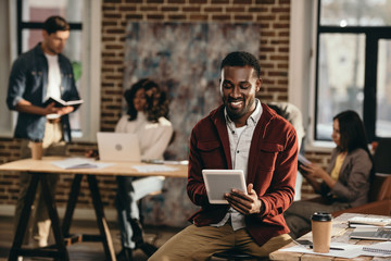 smiling african american casual businessman holding tablet with colleagues working behind in loft office