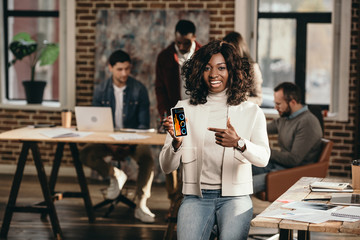 african american casual businesswoman pointing at smartphone with graph on screen in loft office with colleagues behind