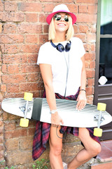Beautiful hipster girl with skate board wearing sunglasses leaning on the brick wall