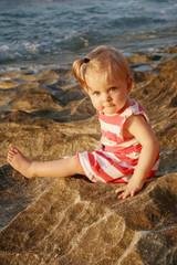 One-year-old baby girl in a red and white dress sitting on the rocky coast, spending time outdoors is important for little ones