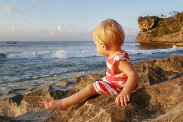 Fototapeta na wymiar One-year-old baby girl in a red and white dress sitting on the rocky coast, spending time outdoors is important for little ones