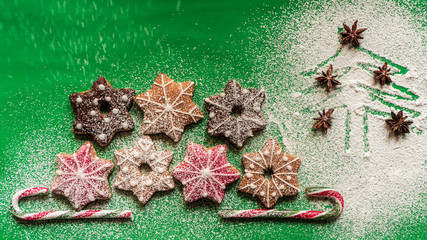 Homemade gingerbread cookies and candy canes on a green table sprinkled with icing sugar next to a Christmas tree drawing, flat lay. Winter holidays concept. 
