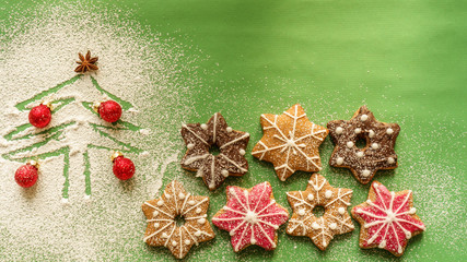 Homemade gingerbread cookies of different colors decorated on green background next to a Christmas tree drawn in icing sugar with red baubles. Winter holidays concept.