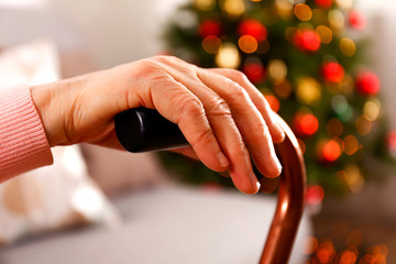 Elderly woman celebrating Christmas at home, with decorated holiday pine tree on background. Old lady at nursing home. Close up, copy space, cropped shot.