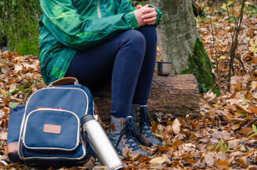 Fototapeta premium Young woman in a green jacket and black jeans drinking tea from a cup or mug in the autumn forest. Walk in the woods. Stop for a rest and a snack. Hiking in the fall.