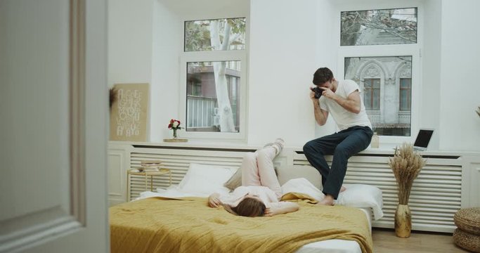 Young guy make pictures for his girlfriend in their cozy bedroom , she is laying down on the bed and wearing a cute pink pajamas. 4k
