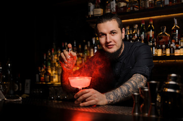 Professional young bartender sprinkling red coclored bitter into a cocktail glass