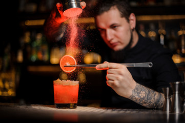Fototapeta na wymiar Tattooed professional bartender adding spices powder into a cocktail glass with slice of lemon