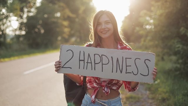 Young beautiful woman hitchhiking standing on the road holding 2 happiness sign. Summer time
