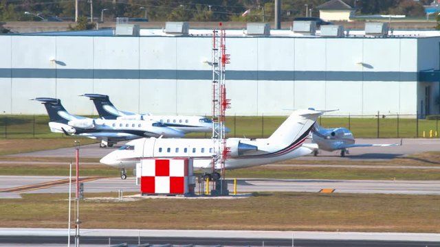 Generic Private Aviation Scene at Airport with White White Executive Business Jet Moving on Airport Ground Taxiways with unmarked Airplanes and Hangars in the Background on a Sunny Day