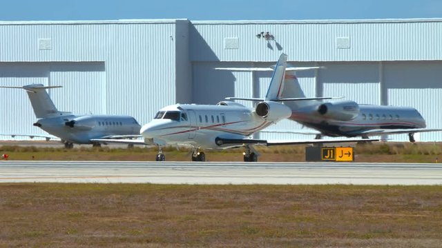 Generic Business Jet Turning onto Airport Runway on a Sunny Day with Private Aviation Hangars and Parked Executive Airplanes in the Background