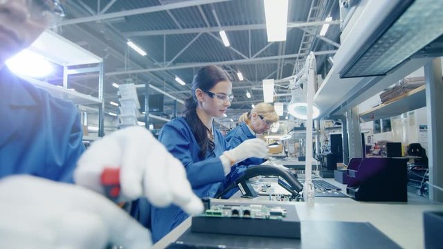 Female Electronics Factory Worker in Blue Work Coat and Protective Glasses is Assembling Laptop's Motherboard with Tweezers. High Tech Factory Facility with Multiple Employees. 