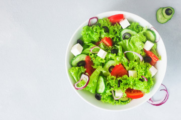 Greek salad in white bowl on grey light background with copy space - lettuce, feta, tomatoes, red onion, cucmber and olives, served with lemon and olive oil