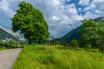 Fototapeta na wymiar Wegkreuz und Bank unter einem Baum strümisches Wetter in den Alpen