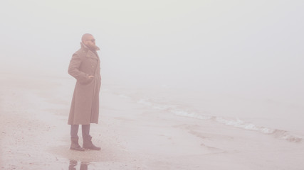 Solitary man with beard and greatcoat is standing by the sea (on the beach) with calm sea and fog...