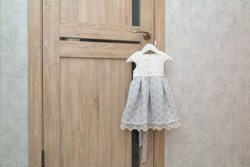 Beautiful festive baby dress hanging on door handle near grey wall. Preparing for the holiday. Waiting for birthday or new year, Christmas celebration. Gift for a girl. Childhood, matinee, happiness.